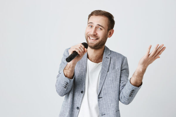 Talented male model wearing gray stylish jacket holding microphone and singing songs while posing against gray background. Bearded singer smiling joyfully